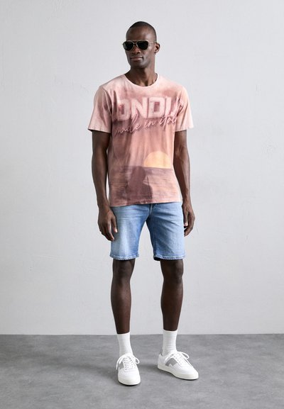 Camiseta de manga corta con un diseño degradado de rosa a durazno, que presenta un gráfico de atardecer y el texto "ONDUI made in Italy". Combinada con shorts de mezclilla.