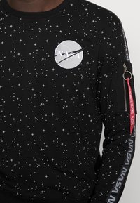 Svart sweatshirt med vita stjärnmönster, NASA-logotyp på bröstet, ficka med dragkedja och röd tagg "REMOVE BEFORE FLIGHT" på ärmen med NASA-textstripe.