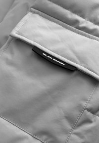 Grijze geïsoleerde stof met een gewatteerde textuur heeft een klepzak. Een zwart label met "BLOCK PAVERS" is opvallend bevestigd.