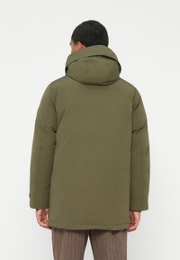 Olíva zöld párnázott téli dzsekis férfi, nagy kapucnival, hátulról nézve egy sima világos háttér előtt.