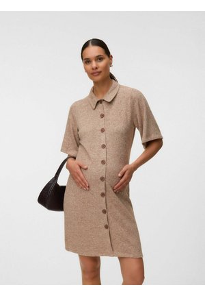 Donna con i capelli scuri raccolti in una coda bassa che indossa un vestito beige con bottoni e maniche corte, che tiene una borsa nera sulla spalla su sfondo bianco.