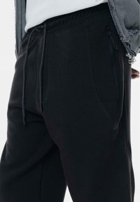 Pantaloni de trening negri cu talie elastică și șnururi ajustabile. Au buzunare laterale cu fermoar și un material neted, texturat.
