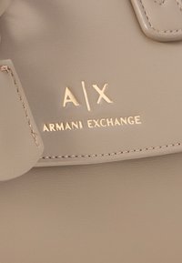 Borsa a mano in pelle beige con logo dorato in rilievo "A|X ARMANI EXCHANGE", caratterizzata da dettagli cuciti e una texture liscia.