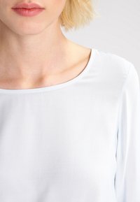 Blusa azzurra chiara a maniche lunghe con scollatura tonda. Tessuto di consistenza liscia, con un design semplice senza motivi o decorazioni.