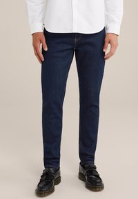 Sötétkék farmernadrág, slim fit szabású, klasszikus öt zsebes dizájnnal és varrott részletekkel. Fekete loafers cipővel párosítva.