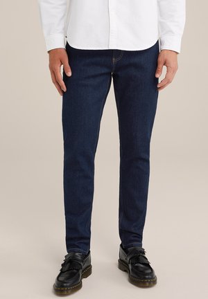Jeans de mezclilla azul oscuro con un corte ajustado, que presentan un diseño clásico de cinco bolsillos y detalles de costura. Combinados con mocasines negros.