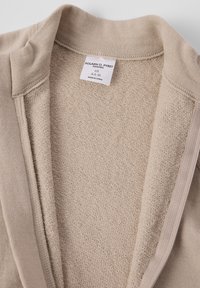 Beige textiel met een open kraag en een zichtbaar wit kledinglabel met de tekst "Polarn O. Pyret Sweden 68 Made in China."