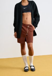 Svart fluffig cardigan, grå sportbh, brun kjol med dragkedja framtill, vita knähöga strumpor, svarta skor med spänne, står på ett gult golv.