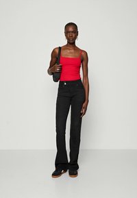 Top de tirantes rojo ajustado, Jeans negros de campana y zapatillas negras. La modelo lleva un bolso negro texturizado. Fondo simple de color neutro.