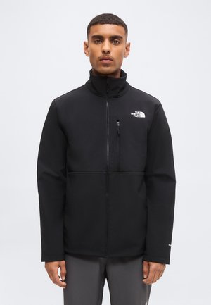 Man wearing a black zip-up North Face jacket with a high collar, standing against a plain light background.