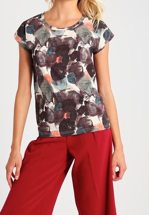 Frau trägt ein kurzärmeliges Oberteil mit buntem abstraktem Muster und hochtaillierte, weit geschnittene rote Hose vor einfarbigem Hintergrund.