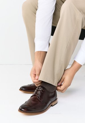 Personne portant un pantalon beige et une chemise blanche ajuste la jambe du pantalon par-dessus une chaussure habillée en cuir brun foncé avec des détails brogue.