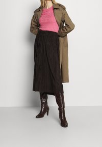 Cappotto trench marrone, maglione rosa a coste, gonna midi plissettata nera e stivali lucidi marrone al ginocchio. Focalizzarsi sulla texture e sulle sovrapposizioni nello styling.