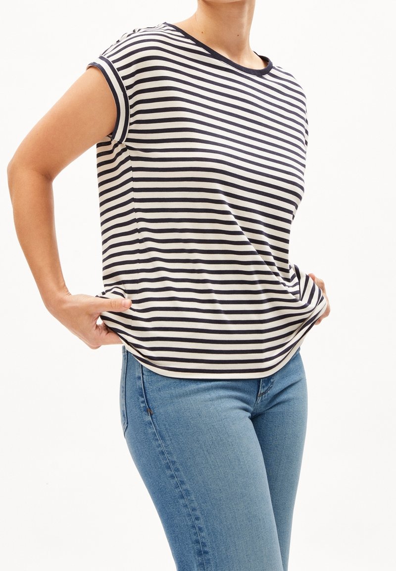 Persona con una camiseta de manga corta a rayas blancas y negras y jeans azules, sujetando los bordes de la camisa cerca de la cintura.