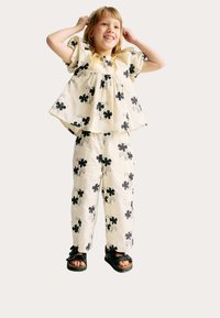 Tenue crème claire composée d'un top à manches courtes amples et froncées, accompagné d'un pantalon large, présentant un motif floral noir. Associée à des sandales noires.