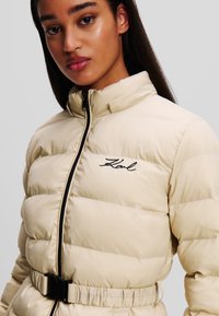 Beige pufferjack met een hoge kraag, zwarte rits en een ceintuur in de taille; heeft gestikte panelen en een geborduurd logo op de borst.