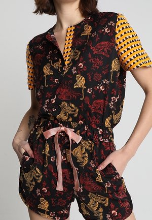 Kobieta ubrana w czarny kombinezon z złoto-czerwonymi wzorami zwierzęcymi, pomarańczowymi geometrycznymi krótkimi rękawami i różowym ściągaczem w pasie.