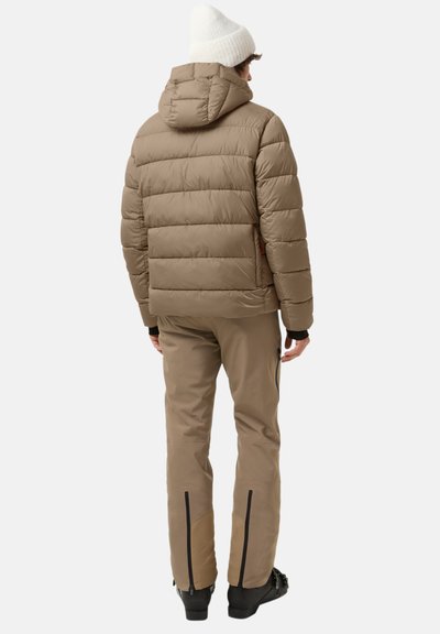 Brown puffer jacket with horizontal stitching, hood, and side pockets, paired with tan pants featuring zippers at the ankles and black footwear.