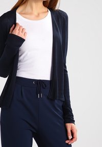 Navy blue cardigan made of lightweight fabric, open front with long sleeves. Worn over a white t-shirt and paired with dark blue elastic waist pants.