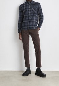 Marine geruite button-up shirt met witte lijnen, enkele borstzak, bruine slim-fit broek en zwarte enkellaarzen met dikke zolen.