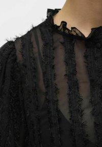 Mustmust tšifonist blouse, millel on volangkaelus, vertikaalsed triibud ja friteeritud aktsendid. Tekstureeritud kangas kerge välimusega.