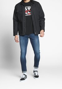 Svart knappjacka med två framfickor, bärs över en svart grafisk t-shirt. Ljust blå slim-fit jeans och svarta sneakers med vita detaljer.