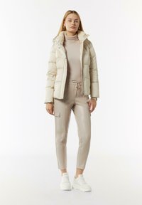 Helle beige Pufferjacke mit einem mit Fell gefütterten Kragen; passender beiger Rollkragenpullover; schmal geschnittene beige Kunstlederhosen; weiße Sneakers.