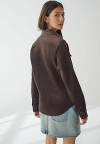 Brun kordfløjs skjorte med knapmanchetter, der har en rygskulder og rund kant, parret med en lyseblå denimnederdel.