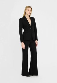 Černé šité sako s hlubokým výstřihem do V a jednoknoflíkovým zapínáním, ve spojení se zvonovými černými kalhotami, oba z hladké látky.