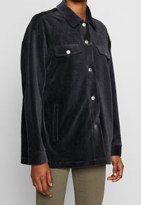 Chaqueta de pana negra con cuello, botones plateados, dos bolsillos en el pecho y un corte relajado, que presenta una textura suave y mangas rectas.