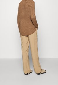 Brauner Strickpullover mit abgerundetem Saum, kombiniert mit locker sitzenden beigen Hosen und flachen schwarzen Sandalen, von hinten betrachtet.