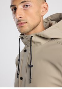 Beige waterproof jacket with a high collar, black drawstrings, and snap buttons; smooth texture and minimalistic design.