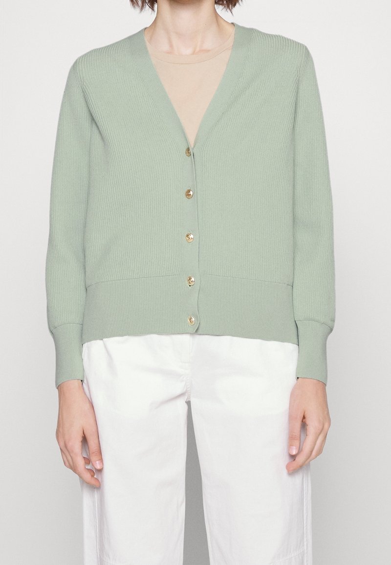 Cardigan vert clair à col en V avec boutons, porté par-dessus un haut beige, associé à un pantalon blanc ample sur une personne debout.