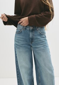 Svetlo modri široki kavbojke z visokim pasom, opremljene z petimi žepi in rahlimi izbliski, v kombinaciji z rjavim dolgim rokavom.