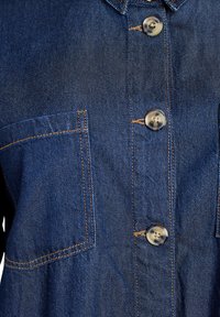 Traper košulja u tamnoplavoj nijansi, s narančastim šavovima, dva prednja džepa i kopčanjem na gumbi s prozirnim plastičnim gumbima.