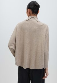 Frau mit dunklen Haaren zu einem Dutt gebunden, trägt einen lockeren beigen Pullover und schwarze Hose, von hinten gegen einen einfachen hellen Hintergrund blickend.