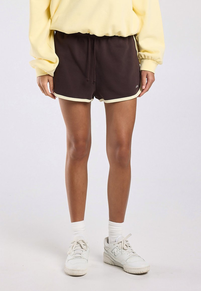 Person trägt dunkelbraune Sportshorts mit hellem Rand, ein gelbes Sweatshirt, weiße Socken und weiße Turnschuhe und steht vor einem einfarbigen Hintergrund.