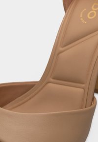 Gros plan sur la semelle intérieure rembourrée et la lanière supérieure beige d'une chaussure à talons hauts, mettant en valeur sa texture lisse et son design minimaliste.