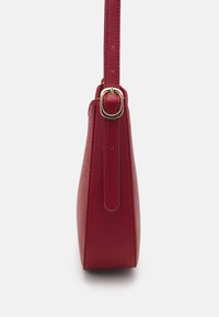 Punainen nahkainen käsilaukku, jossa on jämäkkä muoto, sileä pinta ja pitkä hihna, joka on kiinnitetty kultaisen ovalin muotoisen soljen avulla.