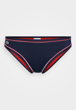 Tengerészkék bikini alsó piros és fehér díszítéssel. Alacsony derekú dizájnnal rendelkezik, és egy kis logó részlet található a bal oldalon. Simább textúrájú.