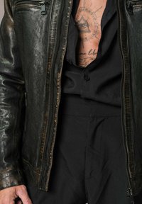 Veste en cuir noir avec une texture usée, dotée d'une fermeture éclair à l'avant et de poches latérales. Portée par-dessus une chemise noire à boutons.