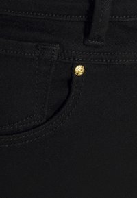 Los jeans negros tienen una vista en primer plano de un bolsillo con herrajes dorados. La tela tiene una textura suave y un detalle de costura visible.