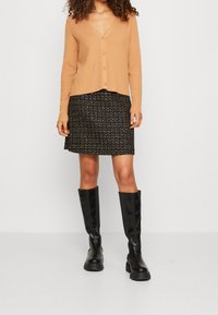 Cárdigan de punto acanalado color camello con escote en V y botones, combinado con una falda de tweed negra con acentos metálicos y botas negras hasta la rodilla.