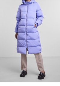 Ljuslila lång pufferjacka med huva, dragkedja fram och sidofickor. Utrustad med quiltat mönster och ribbade muddar. Bärs över beige byxor och svarta skor.