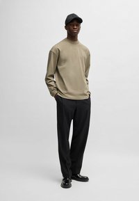 Langärmliges olivgrünes Hemd, schwarze Hose und schwarze Mütze. Das Hemd hat eine lockere Passform und eine glatte Textur. Die Schuhe sind schwarze Slipper mit runder Zehenform.