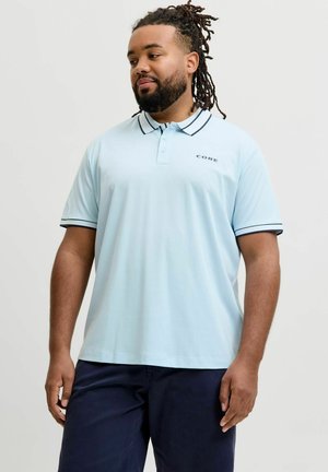 Mężczyzna z dredami i brodą, ubrany w jasnobłękitną koszulkę polo z czarnym wykończeniem i ciemne spodnie, stojący na jednolitym tle.