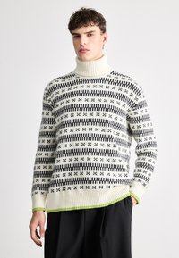 Strikket høyhalsgenser i krem med svart stripete mønster og grønn kant. Har ribbestrikkede mansjetter og kant, laget av mykt stoff.