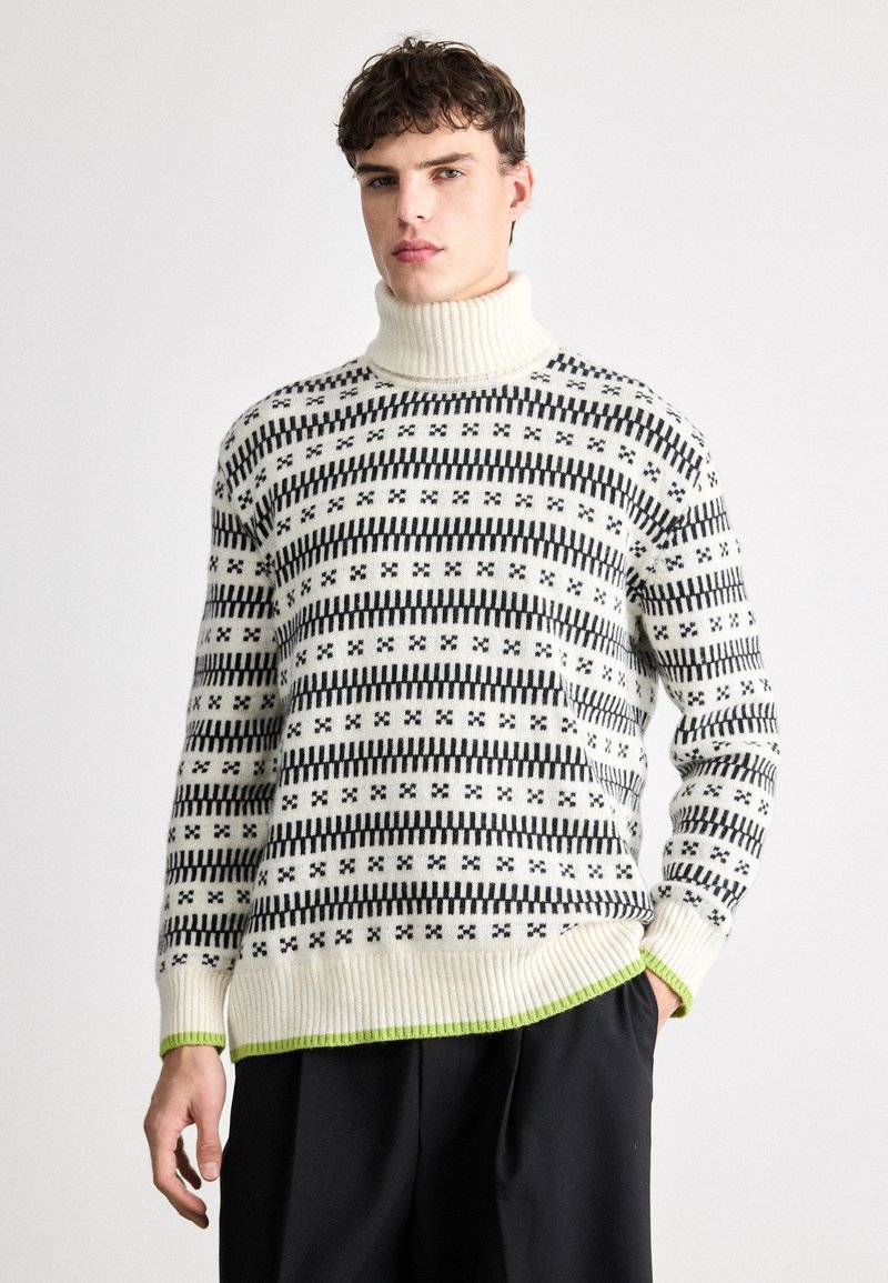 Strikket høyhalsgenser i krem med svart stripete mønster og grønn kant. Har ribbestrikkede mansjetter og kant, laget av mykt stoff.