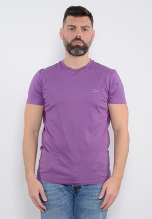 T-shirt viola a maniche corte realizzato in materiale morbido, con scollatura a girocollo e un piccolo logo verde sul lato sinistro del petto, abbinato a jeans azzurri chiari.