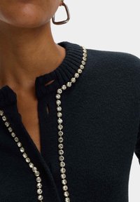 Suéter de punto negro con un cuello acanalado y una línea de cristales transparentes a lo largo de la tapeta de los botones. Tela texturada con cierre de botón.